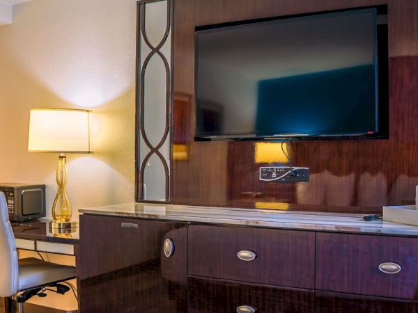 A hotel room with a TV, desk, chair, lamp, and coffee tray on a wooden dresser.
