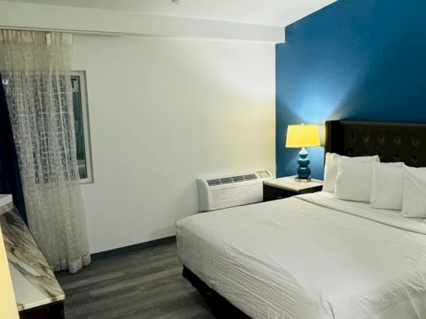The image shows a hotel room with a king-sized bed, blue accent wall, bedside lamps, sheer curtains, and a window beside an air conditioner.