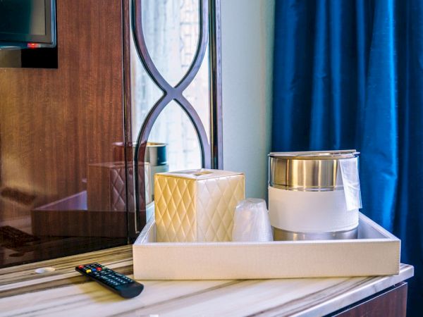 A hotel room's wooden countertop with TV remote, box, ice bucket, glasses, and tray. Blue curtains and window in the background.