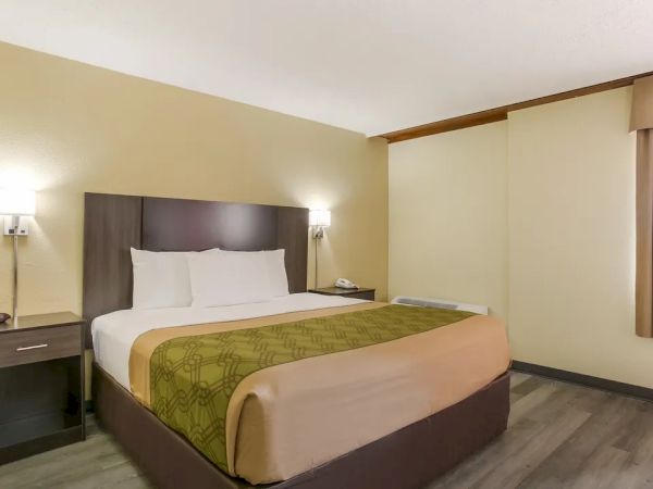 The image shows a neatly arranged hotel room with a bed, bedside tables, and wall-mounted lamps, featuring neutral-colored decor and wood flooring.