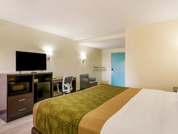 The image shows a hotel room with a bed, TV, desk, chair, microwave, and a blue door. The walls are light-colored with simple decor.