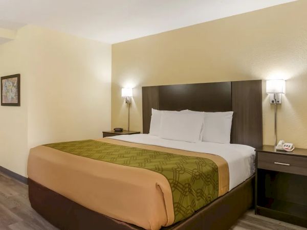 The image shows a hotel room with a neatly made bed, two nightstands, wall lamps, an ironing board, and a picture on the wall.