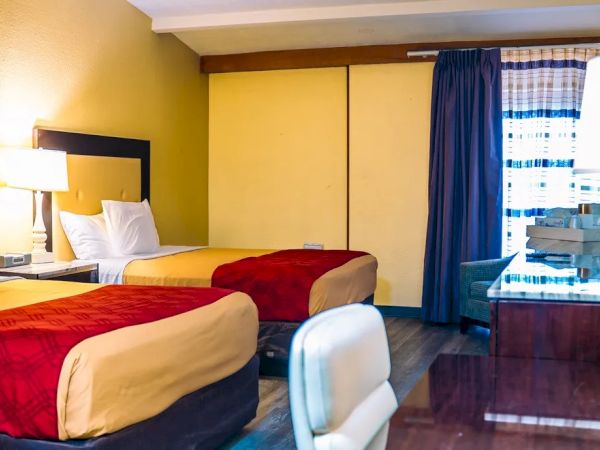 The image shows a hotel room with two beds featuring red and yellow bedspreads, a desk, a chair, and a lamp.