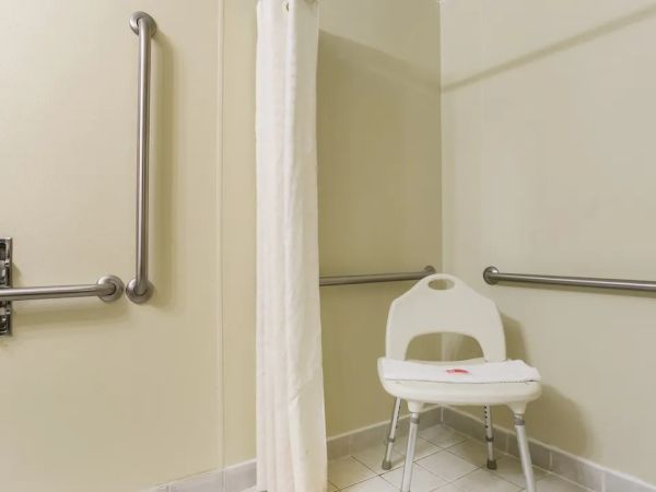 This image shows an accessible shower area with grab bars, a shower curtain, and a white shower chair on a tiled floor.