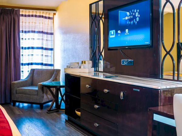 The image shows a hotel room with two beds, a TV on a dresser, a chair, a lamp, and curtains by the window.