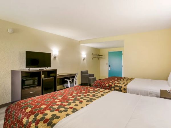 A hotel room with two beds, a TV on a stand, desk with a chair, and a small fridge, designed in warm tones with simple decor.