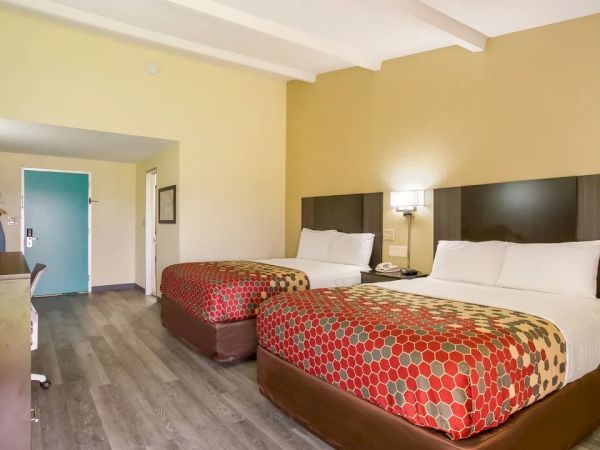 The image shows a hotel room with two double beds, a TV, a desk chair, and a door in the background.