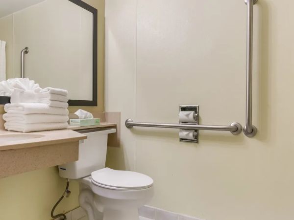 The image shows a bathroom with a toilet, a sink, a mirror, and a stack of towels on the counter.
