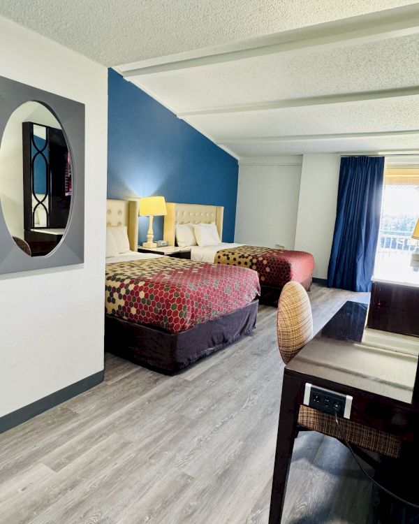 The image shows a hotel room with two beds, a blue accent wall, a desk, and a wall mirror, featuring wood flooring and modern decor.