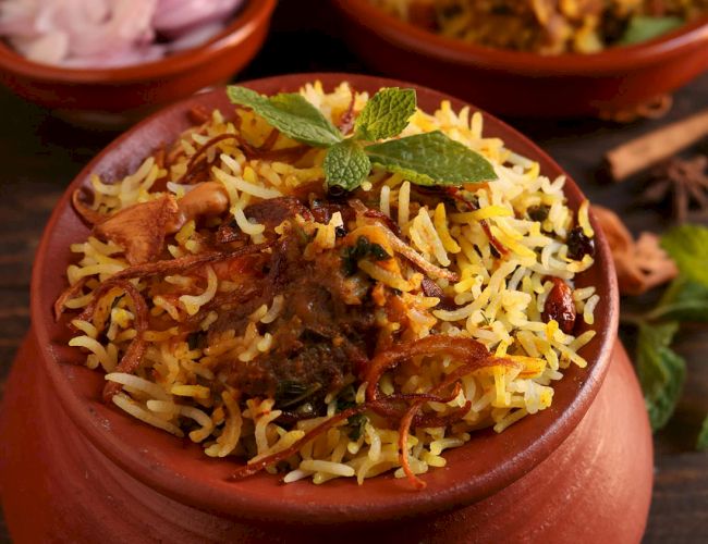 A bowl of biryani garnished with mint leaves is featured, along with a side of sliced onions in the background.
