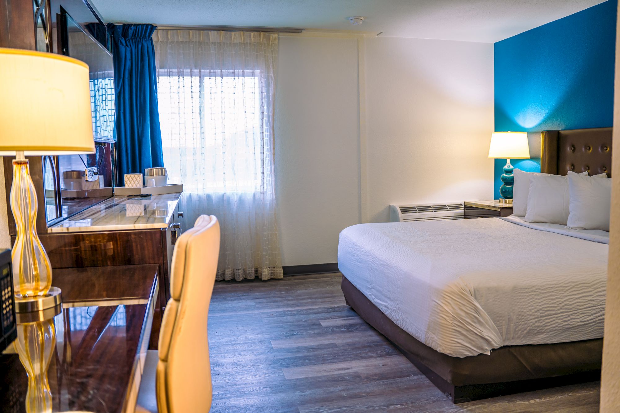 The image shows a modern hotel room with a large bed, a desk, chair, and lamps on both sides, next to a window with curtains.