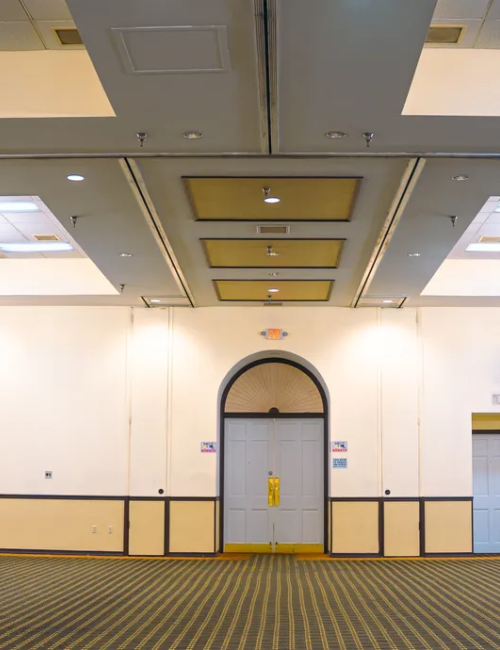 An empty banquet hall with chandeliers, an arched doorway, and a carpeted floor. Stage equipment is visible on the right side of the room.