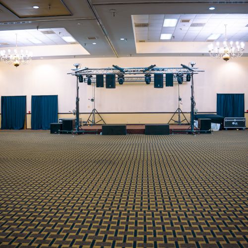 An empty event hall with patterned carpet, chandeliers, and a stage setup with speakers, lighting, and blue curtains in the background.