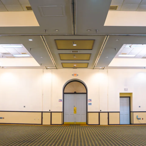 An empty, carpeted ballroom with chandeliers, a high ceiling, and multiple doors, featuring lighting equipment on the right.