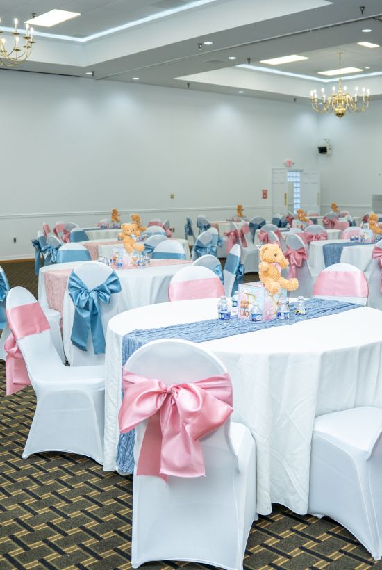 A banquet hall set up for a formal event, with round tables dressed in white cloths, pink and blue chair sashes, and floral centerpieces.