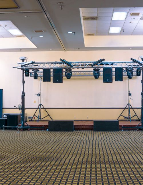 An empty hall with a carpeted floor, stage setup with lighting, speakers, and draped walls; chandeliers hang from the ceiling.