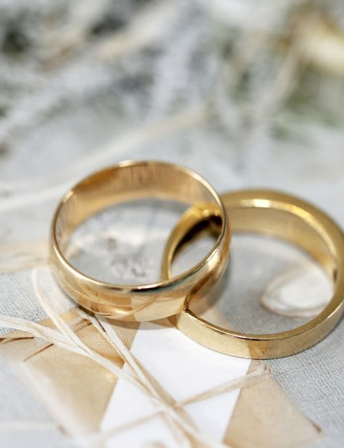 Two gold wedding bands resting on a light, frosted background with delicate white greenery and pale ribbons&mdash;soft, romantic, wedding-themed.