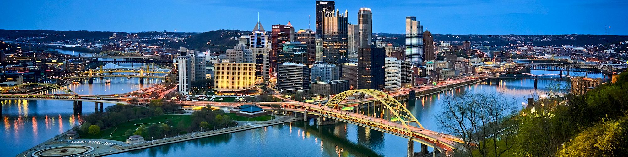 A cityscape at dusk features illuminated bridges, a river, and an incline railway car, with a skyline in the background and lush greenery.