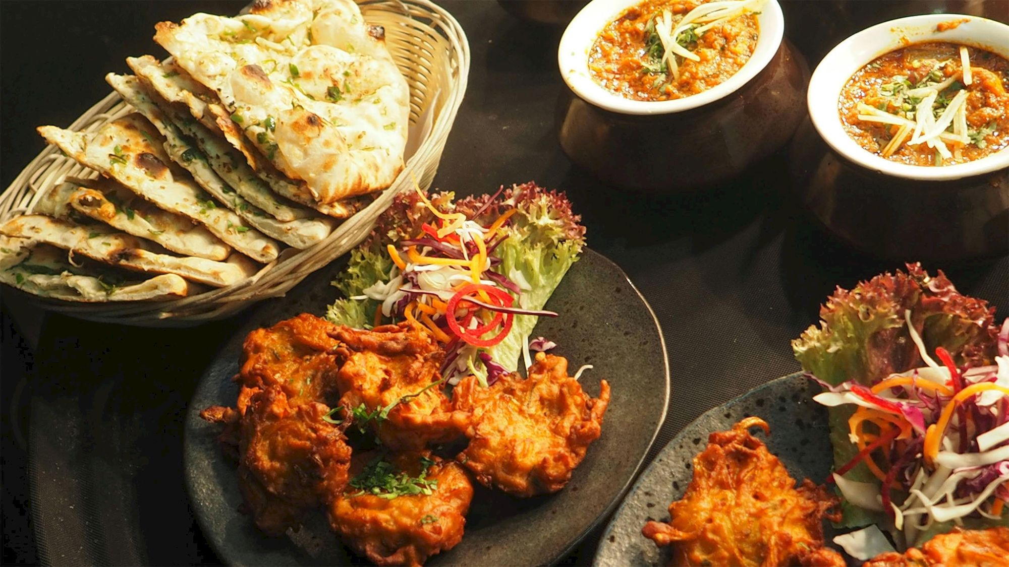 Assorted Indian dishes, including naan, curry in bowls, and vegetable pakoras garnished with herbs and salad on a dark background.