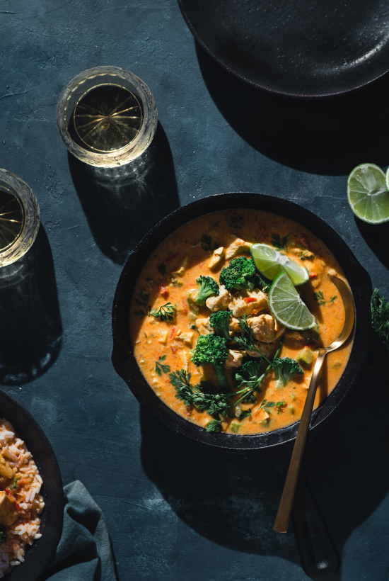 A bowl of curry with lime, broccoli, and herbs, surrounded by drinks, rice, and a dark backdrop; a spoon rests in the bowl.