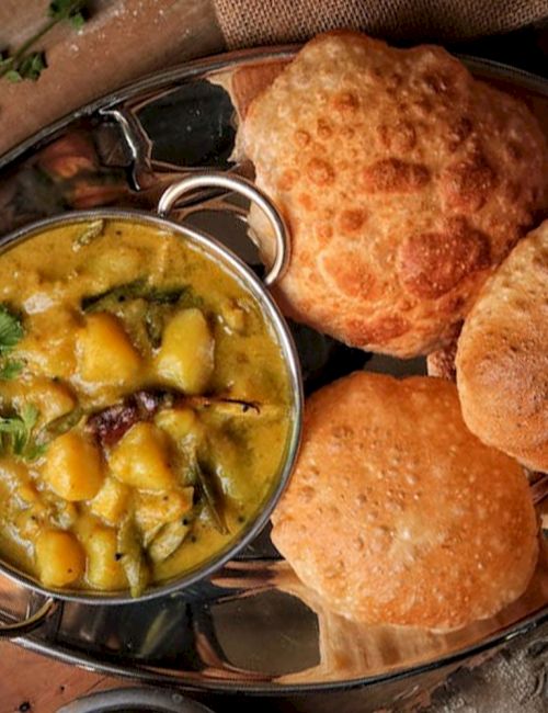 The image shows a dish with puri bread and a bowl of potato curry garnished with cilantro, served on a metal plate.