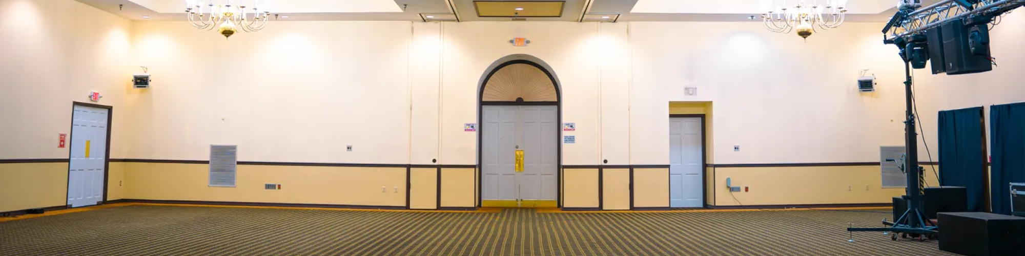 An empty banquet hall with chandeliers, neutral colors, patterned carpet, and lighting equipment near the wall ends the sentence.