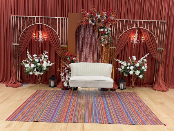 A small stage setup with red curtains, a white sofa, floral arches, two flower arrangements, and a striped rug.