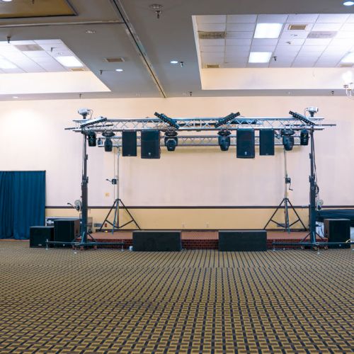 An empty banquet hall with carpet, chandeliers, and a stage setup with lights and speakers.