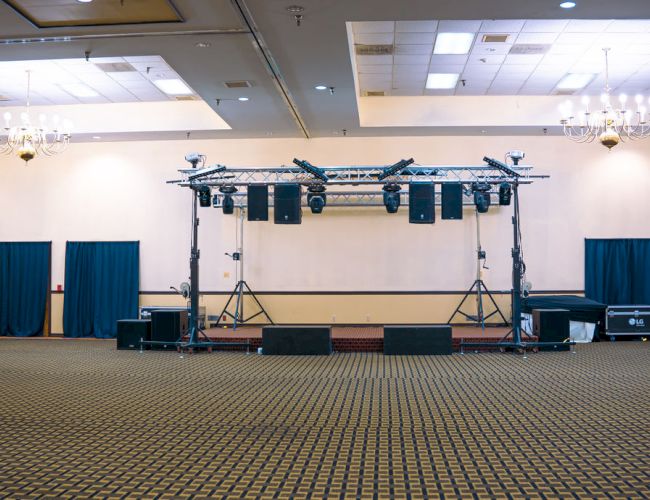 An empty banquet hall with carpet, chandeliers, and a stage setup with lights and speakers.