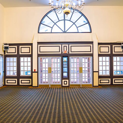 An empty room with patterned carpet, high ceiling, multiple doors, and windows. There are mirrors and wall-mounted lights on the sides.