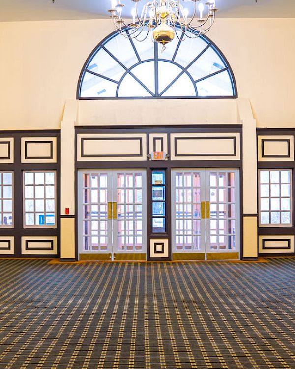 An empty room with patterned carpet, high ceiling, multiple doors, and windows. There are mirrors and wall-mounted lights on the sides.