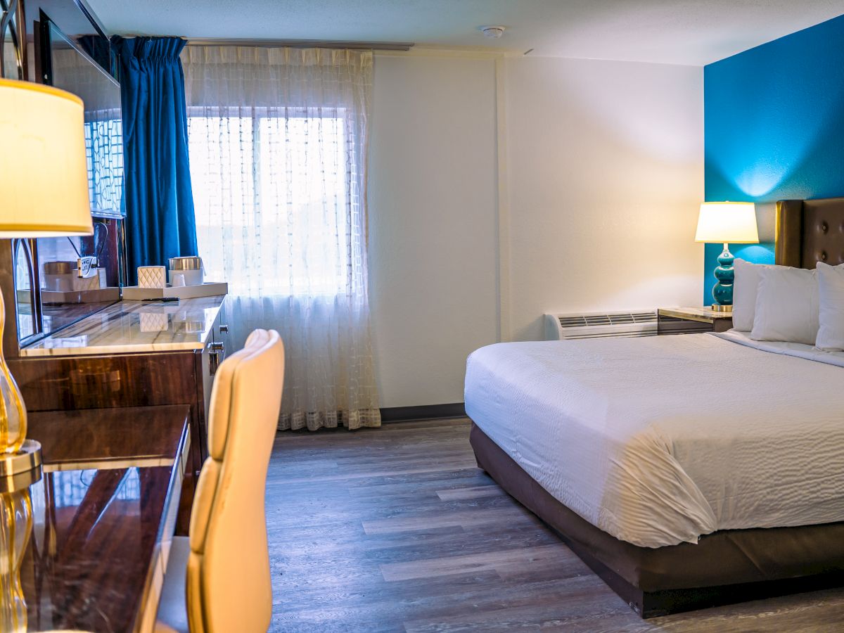 The image shows a well-lit hotel room with a bed, desk, chairs, and two lamps. The decor is modern with a blue accent wall.