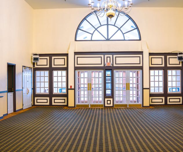 A spacious, empty room with patterned carpet, large arched windows, chandeliers, and mirrors on the walls, featuring a classic design style.