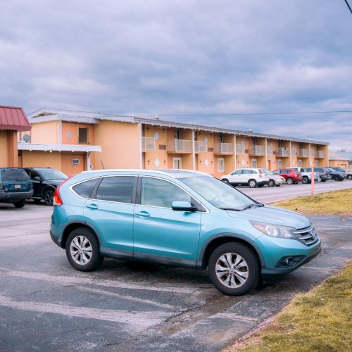 A blue SUV is parked in front of a row of buildings with other cars in a cloudy setting.