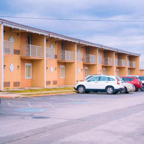 A row of parked cars in front of a two-story building with balconies under a cloudy sky.