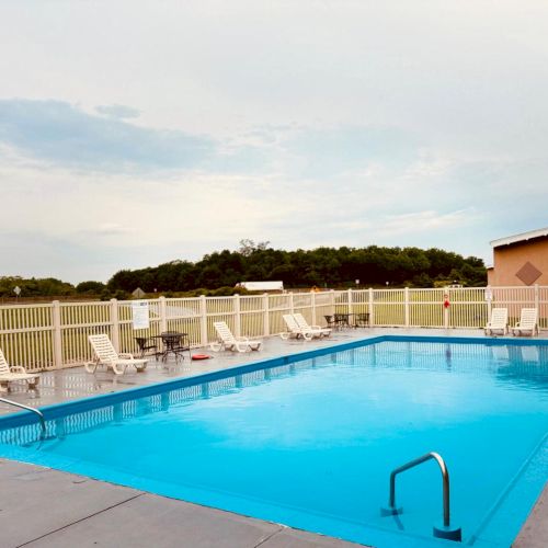 An outdoor swimming pool with lounge chairs and a fence, set against a backdrop of trees and a clear sky.