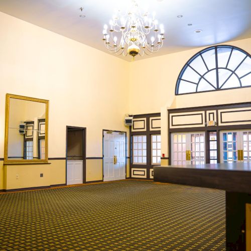 An elegant, spacious room features a large chandelier, arched windows, a mirror, and detailed moldings, with a patterned carpet.