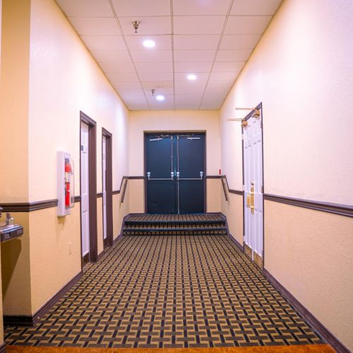 A brightly lit corridor with patterned carpet, doors on the sides, and double doors at the end, featuring ceiling lights and a fire extinguisher.
