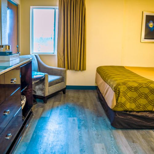 The image shows a hotel room with a bed, a chair, a dresser, a TV, and a window with curtains. The floor is wooden, and there's wall art.