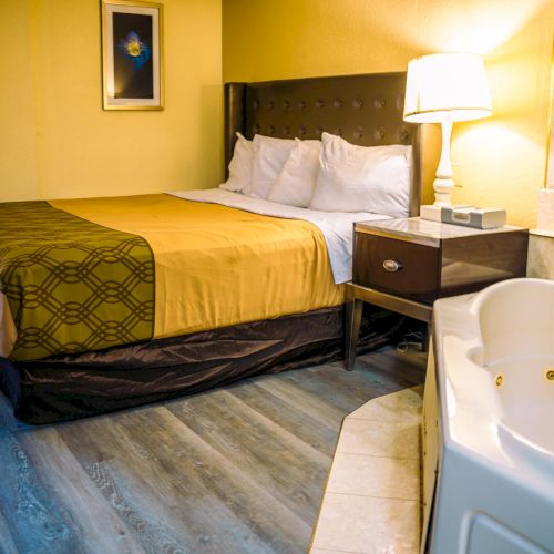 The image shows a hotel room with a bed, nightstand, lamp, and a jacuzzi tub in the corner, featuring wooden flooring.