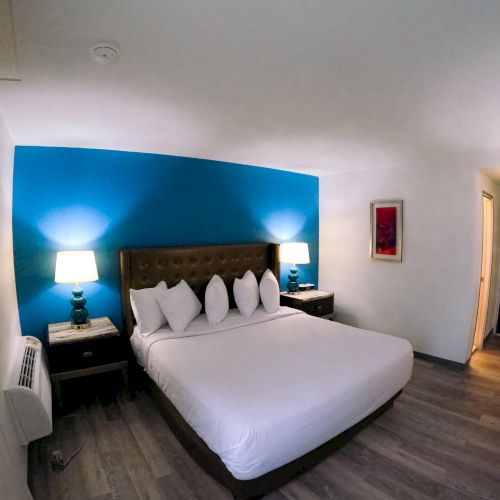 A hotel room with a large bed, two lamps on side tables, blue accent wall, wood flooring, and a desk with a chair.