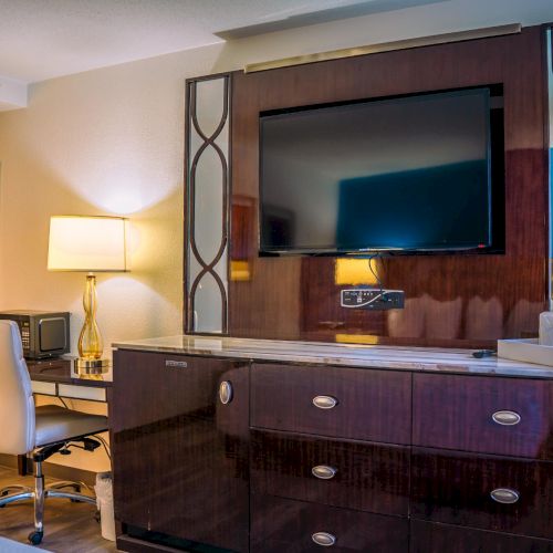 A modern hotel room with a wall-mounted TV, desk, chair, lamp, and decorative mirror on dark wood furniture.