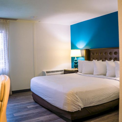 The image shows a neatly made bed in a hotel room with a blue accent wall, a bedside lamp, and a window with sheer curtains.