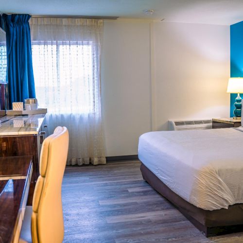 A modern hotel room with a double bed, desk, and chair, featuring warm lighting and a window with sheer curtains.