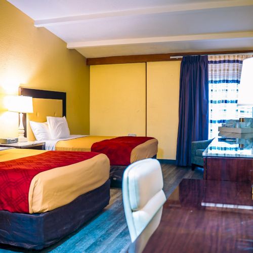 The image shows a hotel room with two double beds, a desk, a chair, a lamp, and a window with curtains.