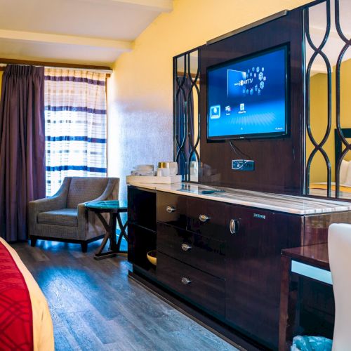 The image shows a hotel room with a TV, two beds, a chair by the window, and a lamp on a dresser. The decor is modern and cozy.