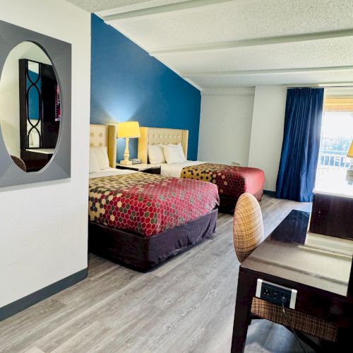 The image shows a hotel room with two beds, a blue accent wall, a desk, chair, and a large window with blue curtains.