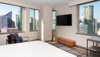 A modern hotel room with a bed, wall-mounted TV, and city view from large windows, showcasing various high-rise buildings.