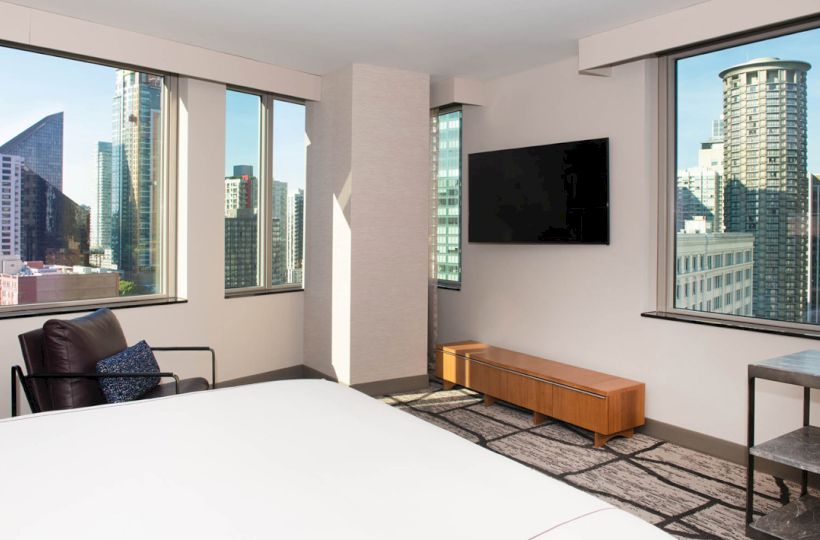 A modern hotel room with a bed, wall-mounted TV, and city view from large windows, showcasing various high-rise buildings.