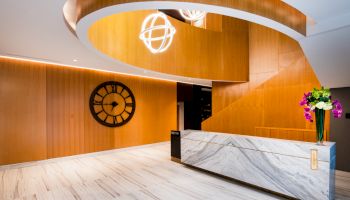 A modern lobby features a marble reception desk, a large wall clock, wooden accents, decorative lights, and a flower vase.